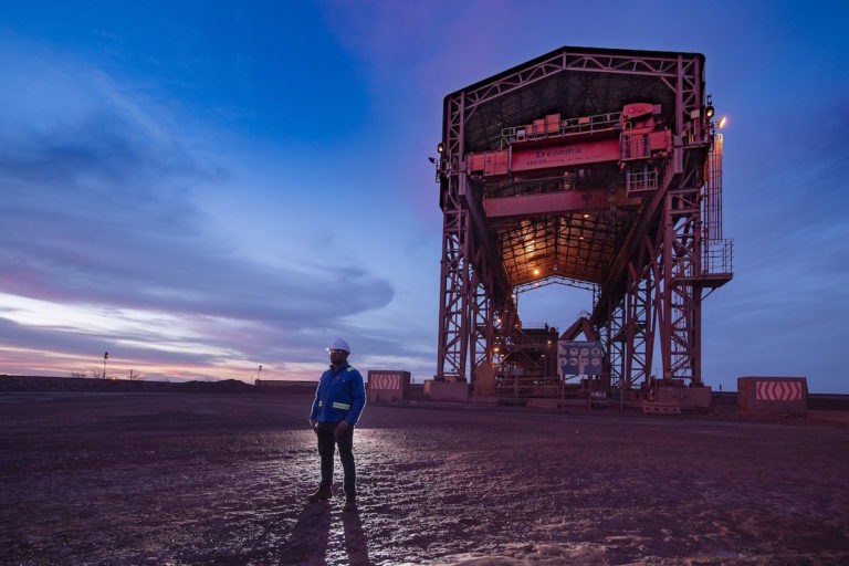 Man in hard hat standing in front of massive primary crusher at sundown at Kolomela iron mine. Image courtesy Anglo-American via Flickr (Fair Use) 